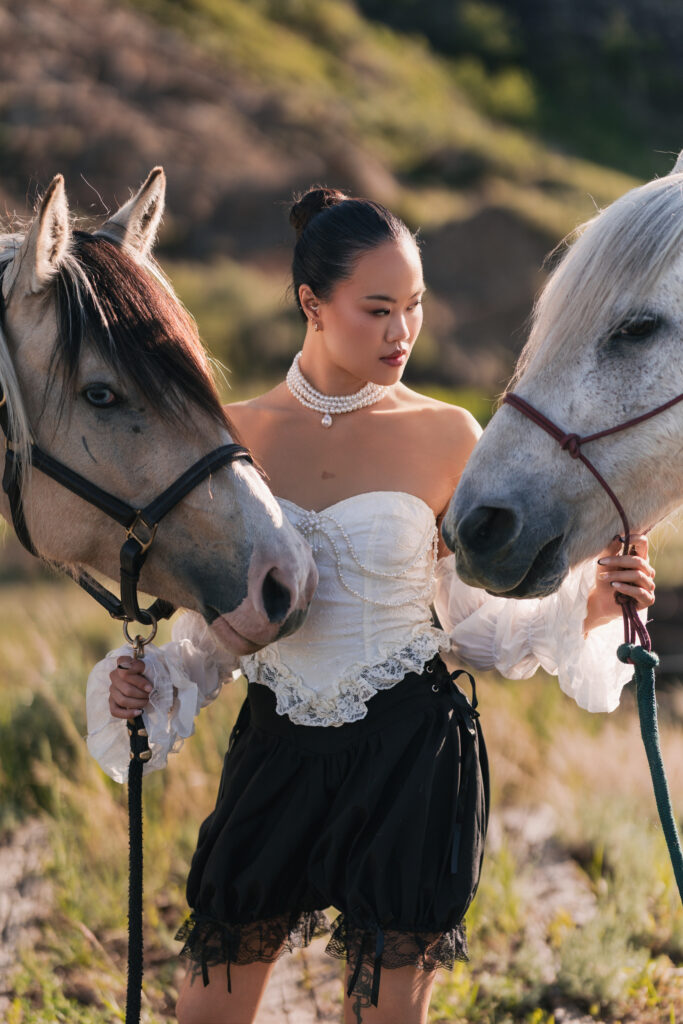 A woman in vintage-style clothing standing outdoors holding the leads of two horses.