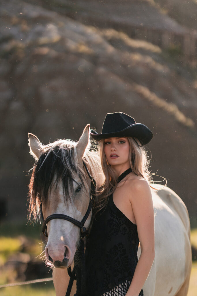 A woman in a black cowboy hat posing closely next to a light-colored horse outdoors.