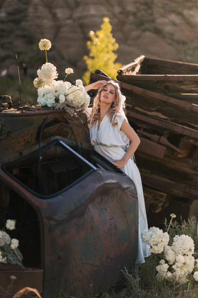 A woman in a long white dress leaning against a rusted vintage vehicle decorated with white flowers.