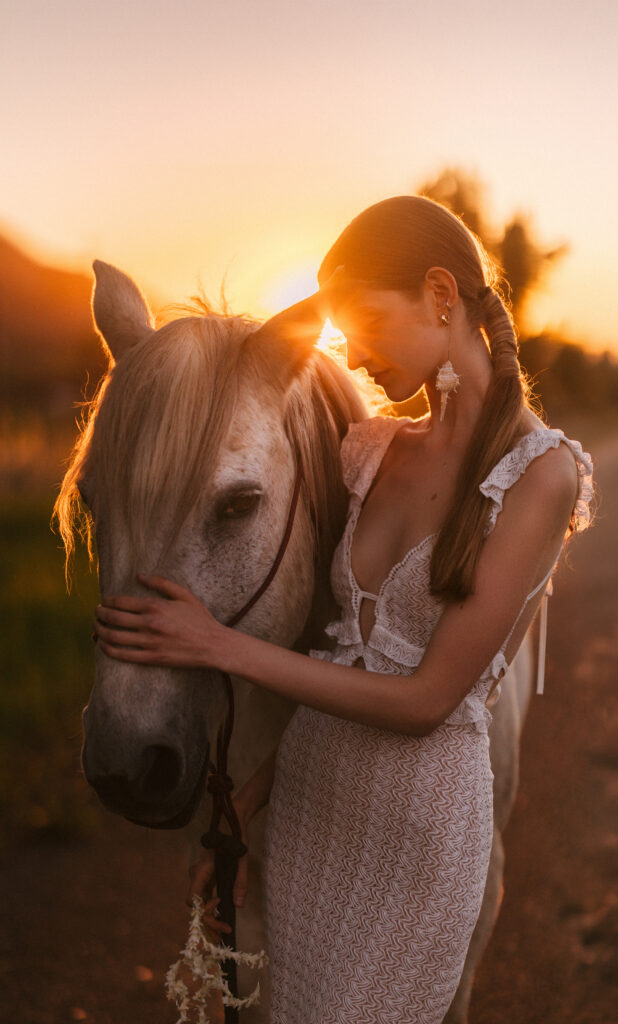 A woman in a white dress gently touching the face of a white horse at sunset.