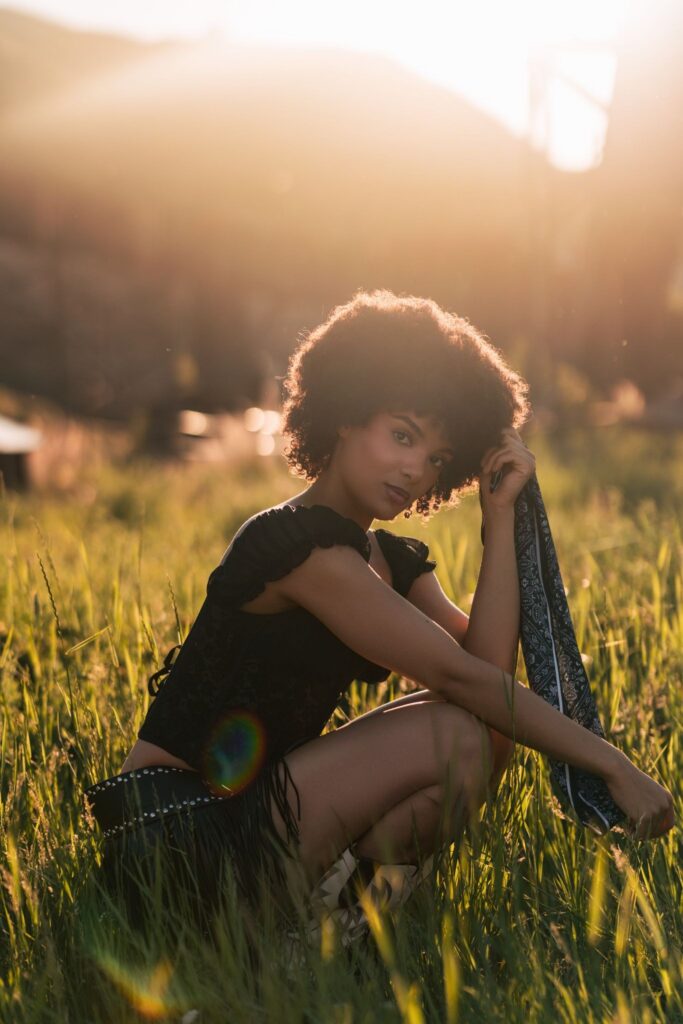 A young woman crouching in a sunlit field of tall grass, holding a dark bandana.
